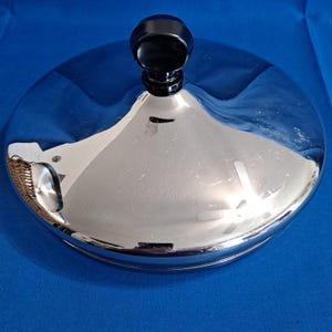 Pode incluir: Uma tampa cromada brilhante com uma pega preta e arredondada. A tampa é circular e reflete o ambiente. A pega está centrada. A tampa é provavelmente feita de metal e destina-se a uma panela ou frigideira. Diâmetro de cerca de 20 cm.