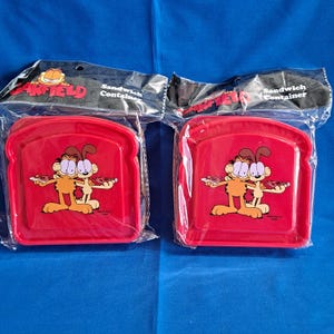 Op de afbeelding: Twee rode Garfield sandwichcontainers in een doorzichtige plastic verpakking. Elke container heeft de vorm van een sneetje brood en heeft een Garfield-afbeelding met pizza. De verpakking leest "Garfield" en "Sandwich Container".