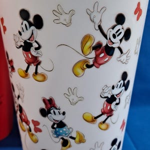Puede incluir: Vaso blanco con ilustraciones de dibujos animados de Mickey Mouse y Minnie Mouse en varias poses. Los personajes est&aacute;n representados con sus atuendos cl&aacute;sicos, con pantalones cortos rojos y zapatos amarillos. El dise&ntilde;o incluye guantes blancos esbozados y lazos rojos.