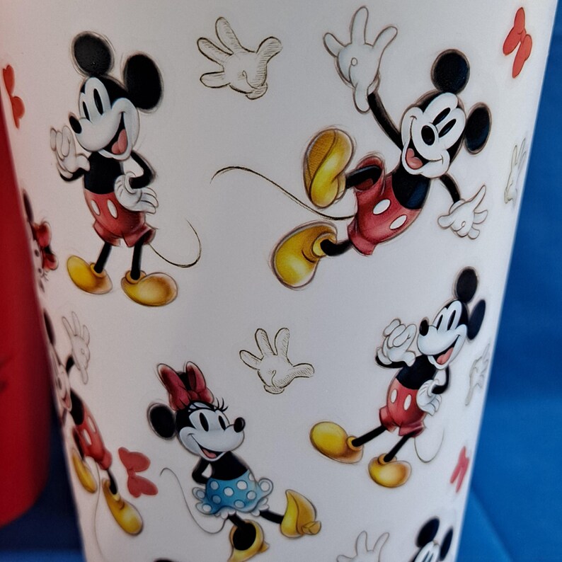 Puede incluir: Vaso blanco con ilustraciones de dibujos animados de Mickey Mouse y Minnie Mouse en varias poses. Los personajes est&aacute;n representados con sus atuendos cl&aacute;sicos, con pantalones cortos rojos y zapatos amarillos. El dise&ntilde;o incluye guantes blancos esbozados y lazos rojos.