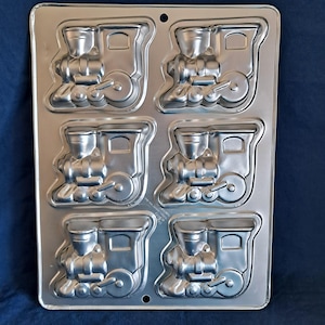 May include: A silver metal baking tray with six train-shaped cavities. Each cavity has a detailed train design, including the engine and wheels. The tray is designed for baking cakes or other treats, and is made of metal.