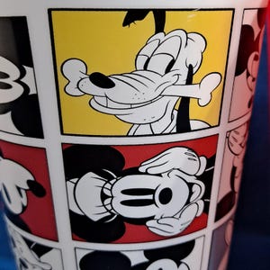 Puede incluir: Vaso blanco con un dise&ntilde;o de c&oacute;mic con personajes de Disney. El vaso muestra a Goofy con un hueso y las manos de Mickey Mouse. El fondo incluye paneles amarillos y rojos, con contornos de personajes en blanco y negro.
