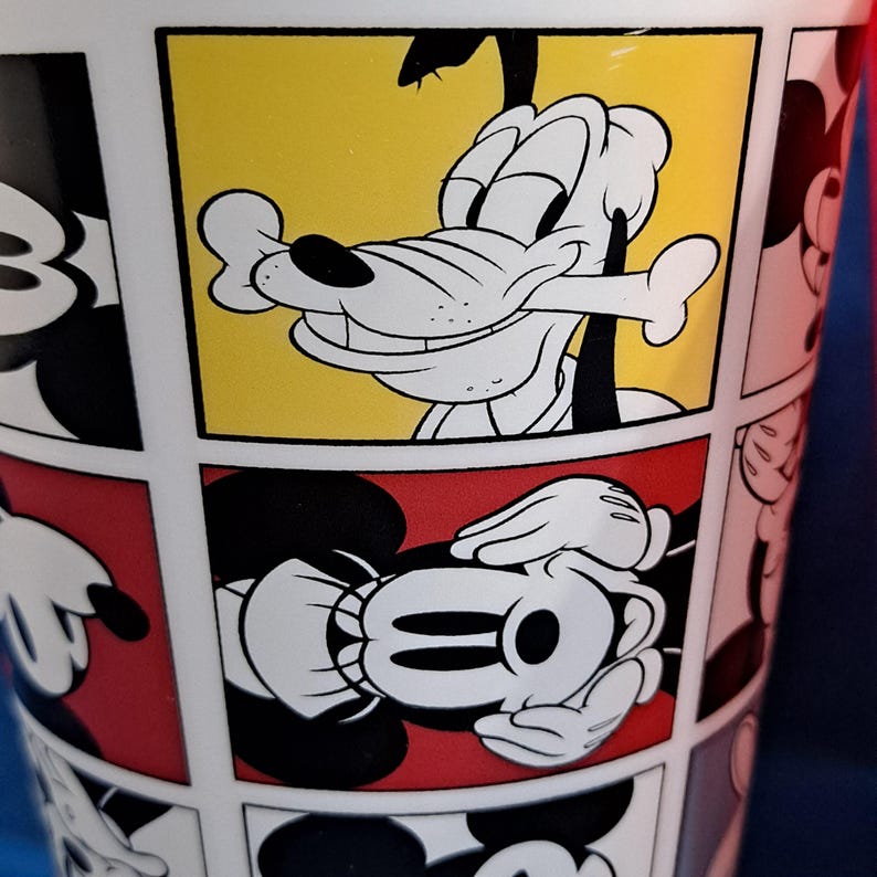 Puede incluir: Vaso blanco con un dise&ntilde;o de c&oacute;mic con personajes de Disney. El vaso muestra a Goofy con un hueso y las manos de Mickey Mouse. El fondo incluye paneles amarillos y rojos, con contornos de personajes en blanco y negro.