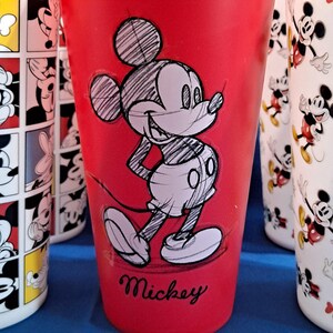 Puede incluir: Vaso de pl&aacute;stico rojo con un boceto de Mickey Mouse. El vaso tiene un dibujo en blanco y negro de Mickey Mouse con la palabra "Mickey" en cursiva en la parte inferior. Otros vasos con dise&ntilde;os de Mickey Mouse son visibles en el fondo.