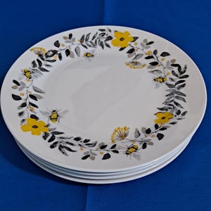 Op de afbeelding: Een stapel van vier witte keramische borden. Elk bord heeft een decoratieve rand van gele bloemen, grijze bladeren en gestileerde bijen. De borden zijn gestapeld op een blauw oppervlak.