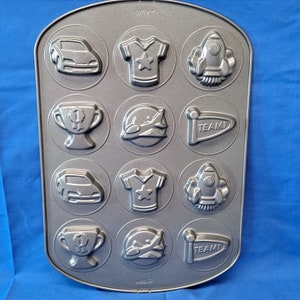 May include: A silver metal baking pan with 12 different shaped molds. The molds are shaped like a car, a rocket, a trophy, a plane, a shirt, and a banner. The banner says "TEAM".