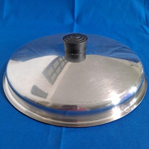 May include: A shiny, stainless steel pot lid with a black, rounded handle. The lid is circular with a slightly raised edge. The surface reflects the surrounding environment. The lid is designed for cooking and food preparation.