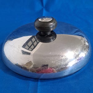 Op de afbeelding: Een glanzende, verchroomde pannendeksel met een zwarte, gegroefde handgreep. De deksel reflecteert de omgeving, inclusief een raam en een persoon. De handgreep heeft een kleine, zilverkleurige bovenkant.