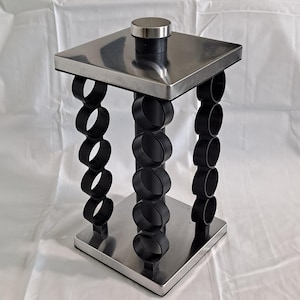 Op de afbeelding: Een moderne kruidenrek met een vierkante roestvrijstalen boven- en onderkant. Het rek heeft zwarte, ringvormige steunen. Bovenop zit een kleine, ronde roestvrijstalen knop. Het ontwerp is minimalistisch en eigentijds.