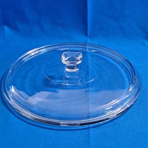 Op de afbeelding: Een helder, rond glazen deksel met een kleine, rechthoekige handgreep. Het deksel heeft een opstaande rand en is ontworpen om op een pot of pan te passen. Het glas is transparant, waardoor de inhoud zichtbaar is. Het deksel is een keukenartikel.
