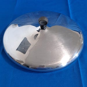 May include: A shiny, silver-colored metal lid with a small, dark knob on top. The lid is round and reflects the surrounding environment. The knob appears to be made of a dark, possibly black, material.