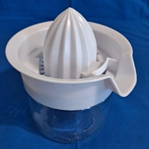 May include: A white citrus juicer with a ribbed reamer, a strainer, and a spout. The juicer sits atop a clear glass jar. The juicer is designed for easy citrus fruit juice extraction.