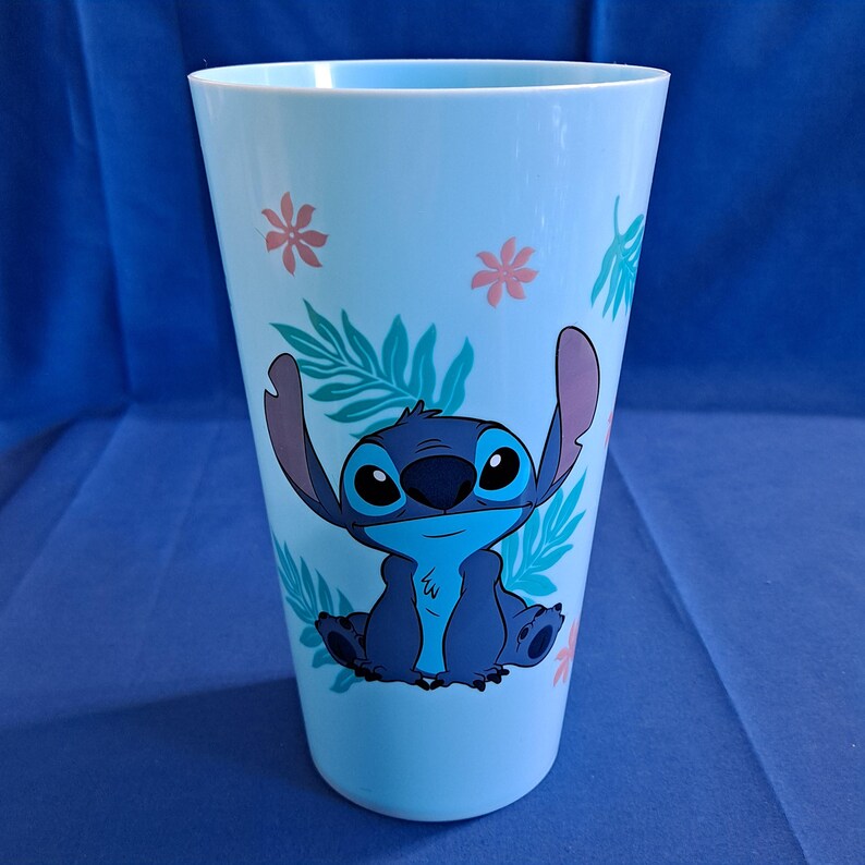 May include: A light blue plastic tumbler featuring a cartoon image of Stitch, with large blue eyes and raised arms. The tumbler is decorated with pink flowers and green leaves. The tumbler is cylindrical and appears to be about 6 inches tall.