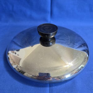 May include: A circular, polished stainless steel pan lid featuring a smooth, reflective surface and a central, black, heat-resistant knob. The lid has a slightly domed shape with a clean, metallic finish, designed to fit securely onto a cooking vessel.