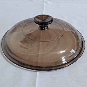 Puede incluir: Una tapa de vidrio transparente teñida de marrón, con un pomo redondo de color marrón oscuro. La tapa es circular, ligeramente abovedada y con borde plateado. Utensilios de cocina.