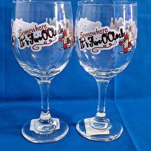May include: Two clear wine glasses with long stems. Each glass has a white sticker with the words "Somewhere It's Five O'Clock" in black and red. The sticker also features illustrations of wine bottles and glasses.