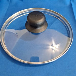 Pode incluir: Uma tampa de vidro transparente com uma alça preta e uma borda prateada. A alça tem o nome da marca "Tramontina" em relevo. A tampa é projetada para utensílios de cozinha.