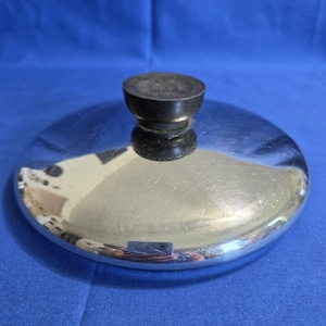 Può includere: Un coperchio circolare per pentola in acciaio inossidabile lucido, caratterizzato da una superficie liscia e riflettente e da un pomello centrale cilindrico di colore marrone scuro. Il coperchio ha una forma leggermente bombata.