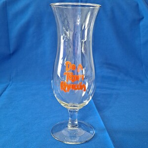 May include: A clear glass hurricane style cocktail glass with a stem and a wide base. The glass has the words "Do a Tropi Kahlua" printed on the side in orange.