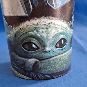 May include: A cylindrical tumbler featuring a detailed image of a character with large eyes and a green complexion. The character's face is the focal point, with a blue and black background. The tumbler is likely made of a durable material.
