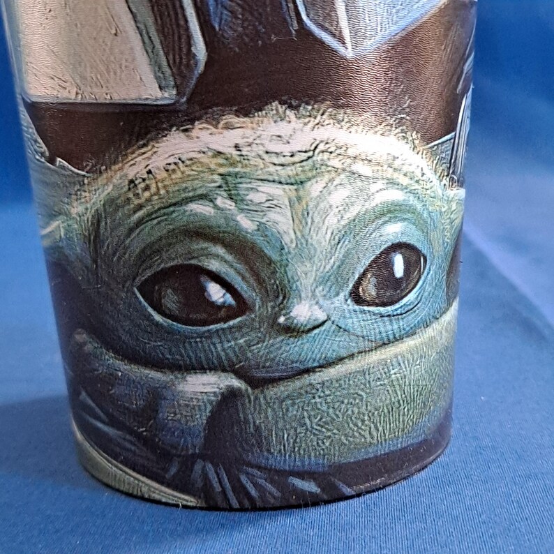 May include: A cylindrical tumbler featuring a detailed image of a character with large eyes and a green complexion. The character's face is the focal point, with a blue and black background. The tumbler is likely made of a durable material.