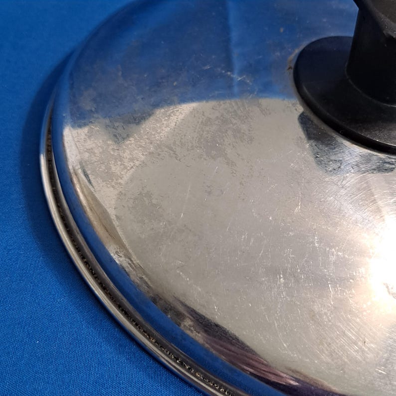 May include: A stainless steel pot lid with a black handle. The lid is round with a shiny, reflective surface and some visible scratches. The edge of the lid has a decorative, textured design.