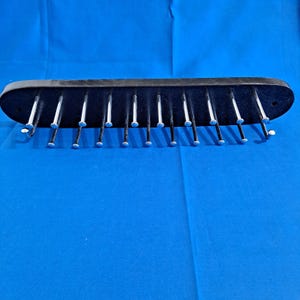 May include: A black tie rack with silver-colored pegs. The rack is made of a dark, possibly plastic material, and is designed to hold multiple ties. The pegs are evenly spaced along the length of the rack.