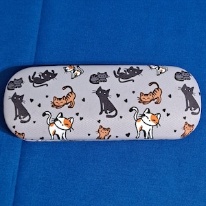 May include: A gray eyeglasses case with a repeating pattern of cartoon cats in various poses and colors, including black, orange, and white. Small black hearts are scattered across the surface. The case is set against a blue background.