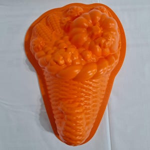 Puede incluir: Molde de silicona naranja con forma de cuerno de la abundancia rebosante de diversas frutas y verduras. El molde presenta texturas detalladas de maíz, uvas y otros productos, perfecto para hornear o manualidades. La cesta tiene una textura tejida.