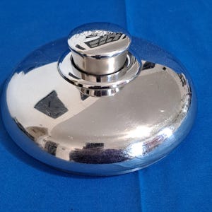 May include: A circular, polished stainless steel pot lid with a reflective surface. The lid features a central, cylindrical knob handle with the brand name MEYER embossed on the top. The design is smooth and domed, showing clear reflections.