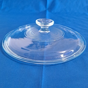 Puede incluir: Una tapa de vidrio transparente con un pomo redondo. La tapa tiene un diseño circular con líneas radiales que emanan del centro. La tapa está sobre una superficie azul.