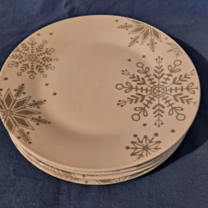 May include: A stack of four off-white ceramic plates. Each plate features a gray snowflake design in various sizes and styles. The plates are stacked on top of each other, showcasing the winter-themed pattern.