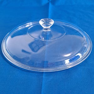 May include: A clear glass lid with a knob handle. The lid is circular and appears to be made of a single piece of glass. The knob is a small, rounded shape. The lid is sitting on a blue surface.