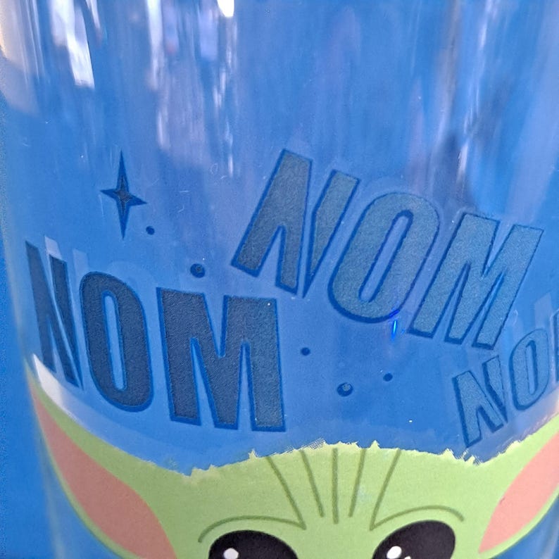 Peut inclure: Un gobelet en plastique bleu avec les mots "NOM NOM NOM" en grandes lettres bleues. Le bas du gobelet pr&eacute;sente un personnage de dessin anim&eacute; vert et beige avec de grands yeux noirs.