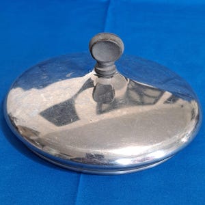 Pode incluir: Uma tampa metálica circular e brilhante com um botão escuro no topo. A tampa reflete o ambiente circundante, e o botão é de cor escura e mate. A tampa é provavelmente para uma panela.