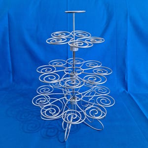 Könnte beinhalten: Ein silberner Cupcake-Ständer aus Metall mit mehreren Etagen. Jede Etage hat dekorative Wirbel zum Halten von Cupcakes. Der Ständer hat eine zentrale Stange und einen Sockel mit gebogenen Füßen. Ideal zum Präsentieren von Backwaren.