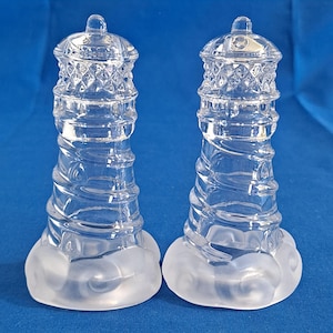 Può includere: Due saliere e pepiere a forma di faro in vetro trasparente. Le saliere e le pepiere hanno un design a spirale sul corpo e una base smerigliata. Le cime sono dettagliate con un motivo a diamante e un piccolo pomello. Lo sfondo è blu.