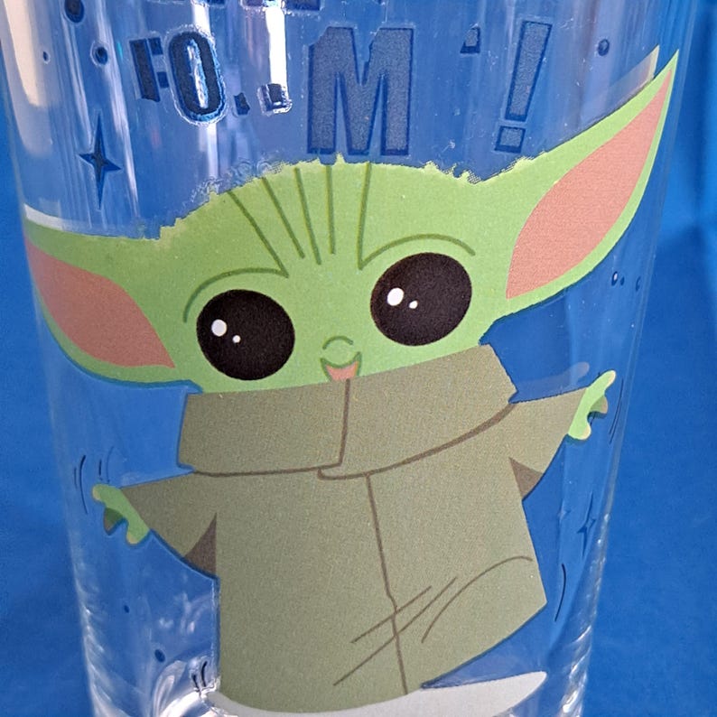 Peut inclure: Gobelet en plastique transparent avec un personnage de dessin anim&eacute; vert avec de grandes oreilles et des yeux noirs. Le personnage porte un manteau beige et a les bras tendus. Le gobelet porte le texte "FO...M!" en bleu.