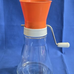 May include: A manual kitchen mill featuring a clear glass base, a white plastic collar, and a bright orange funnel-shaped top. A silver metal crank handle with a white plastic knob is attached to the side for operation of the grinding mechanism.