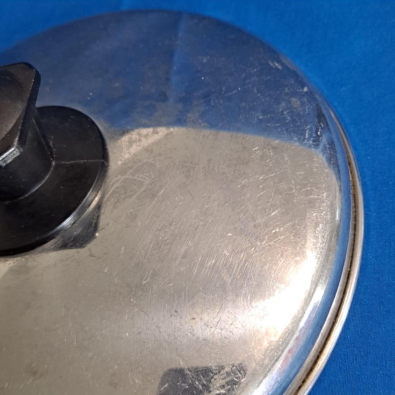May include: Close-up of a stainless steel lid with a black handle. The lid has a shiny, reflective surface with visible scratches. The handle is a dark, matte black. The lid is likely for a pot or pan.