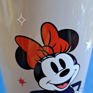 May include: A close-up of a light-coloured mug featuring a smiling Minnie Mouse design. Minnie Mouse has a red bow, black ears, and a black and white face. The mug has star and diamond-shaped accents.