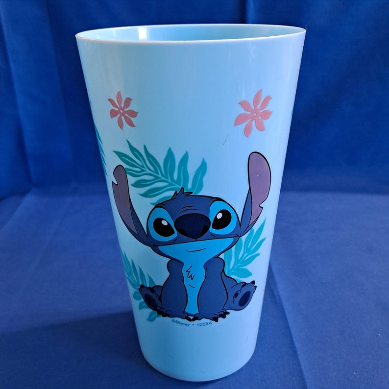 May include: A light blue plastic tumbler featuring a graphic of the Disney character Stitch. The tumbler has a cylindrical shape with a wide opening. The design includes Stitch, tropical leaves, and pink flowers.