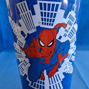 May include: Blue plastic cup featuring Spider-Man in a dynamic pose, with a red and blue costume. The cup has a cityscape design in white, and the Spider-Man graphic is in the center. The cup is cylindrical.