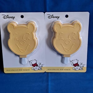 May include: Two yellow silicone ice pop makers featuring Winnie the Pooh's face, packaged on white cards with the Disney logo. Each maker has a light blue stick and a small illustration of Pooh and Piglet.