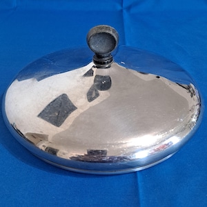 May include: A circular, polished silver-toned metal lid featuring a smooth, reflective surface and a centered, matte black spherical knob handle. The lid has a slightly domed shape and a clean, simple design, providing a functional cover for cookware.