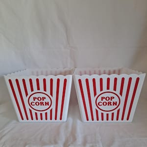 May include: Two white popcorn containers with red vertical stripes and a scalloped top edge. Each container has a red circle with the words "POP CORN" in the center. The containers are empty.