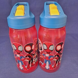 May include: Two red water bottles with blue lids and yellow flip-top spouts. Each bottle features a Spider-Man graphic. The bottles are designed for children and have a straw inside.