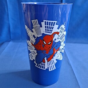 May include: A tall, blue plastic cup featuring a graphic of Spider-Man in a dynamic pose, set against a white cityscape design. The cup is cylindrical and suitable for cold beverages.