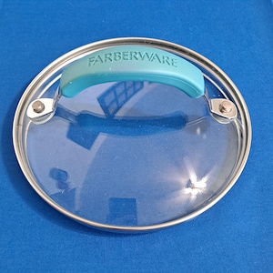 Pode incluir: Tampa de vidro transparente Farberware com aro de aço inoxidável e alça turquesa. A tampa é redonda e possui dois rebites de metal. O logotipo Farberware está impresso.