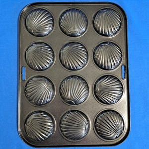 Puede incluir: Una bandeja para hornear metálica con 12 moldes en forma de concha. La bandeja es rectangular con esquinas redondeadas y pequeñas asas. Cada molde mide aproximadamente 7.6 cm de diámetro, ideal para hornear magdalenas o similares.